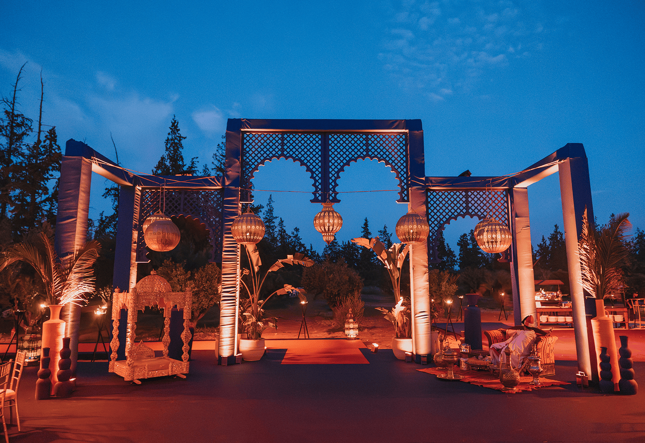 Elegant outdoor gathering with people seated at white tables in front of a historic white building with ornate architecture and potted plants.