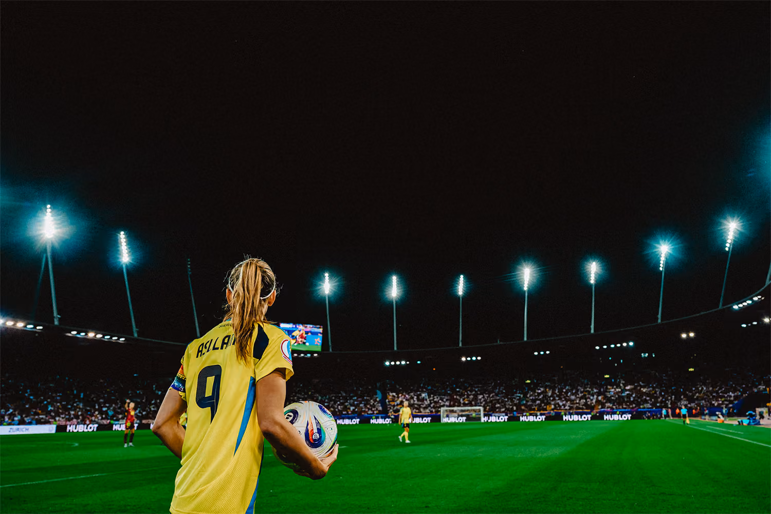 En kvinnlig fotbollsspelare med tröja nummer 9 och namnet Asllani på ryggen håller en fotboll vid sidlinjen på en upplyst stadion under kvällsmatch med publik i bakgrunden.