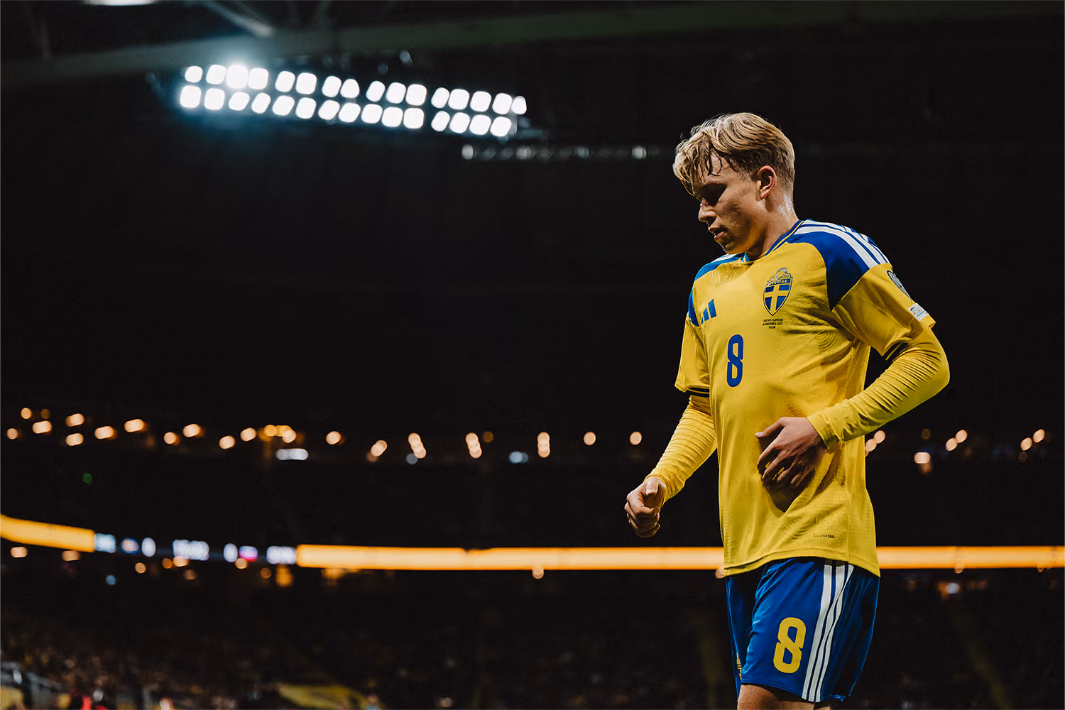 Fotbollsspelare i SVERIGES landslagsuniform med nummer 8, ser nedåt under en match på en upplyst arena med mörk bakgrund.