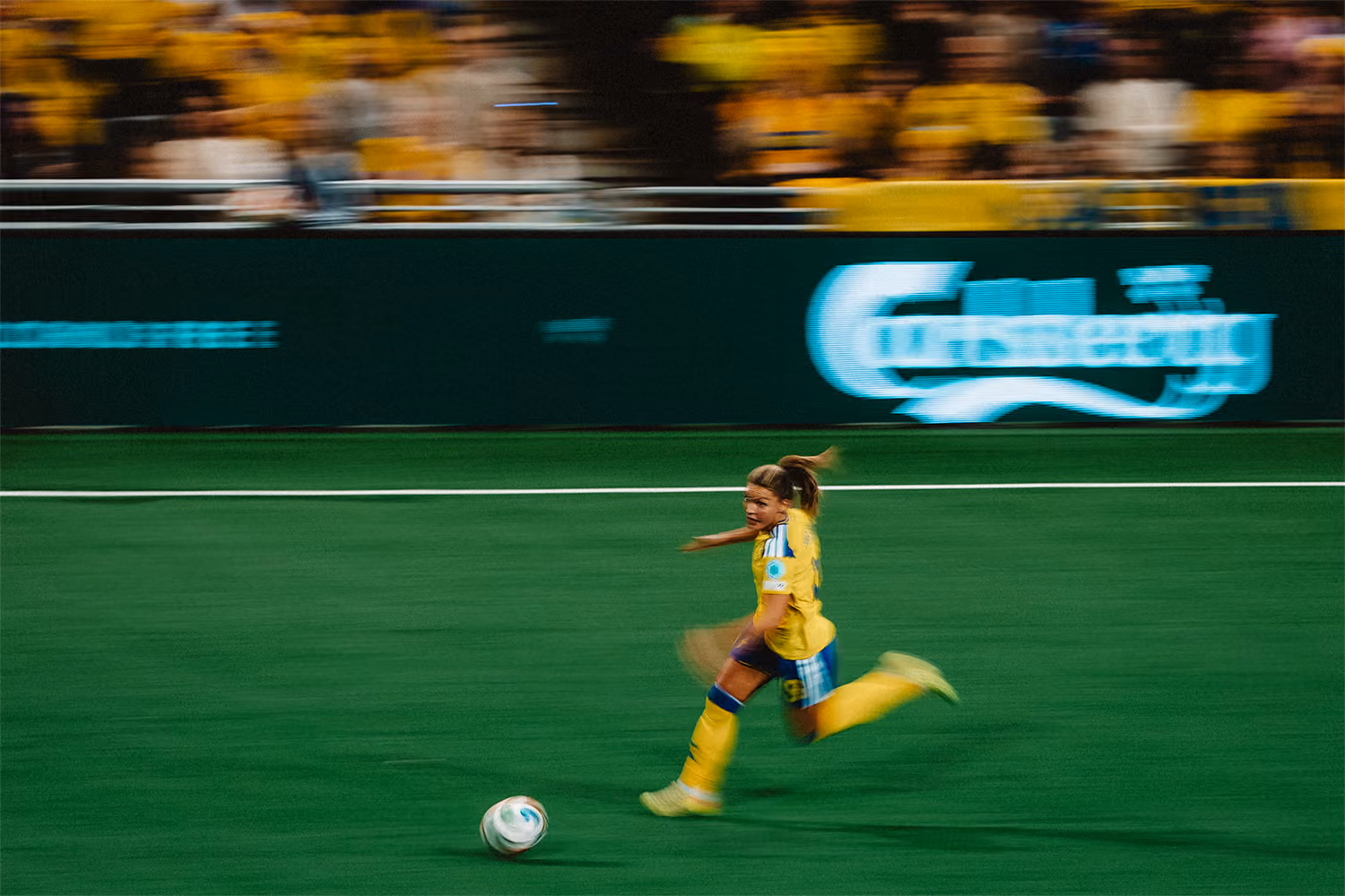 En kvinnlig fotbollsspelare i gul och blå dräkt springer med bollen på gräsplanen under en match, med suddiga åskådare i bakgrunden.