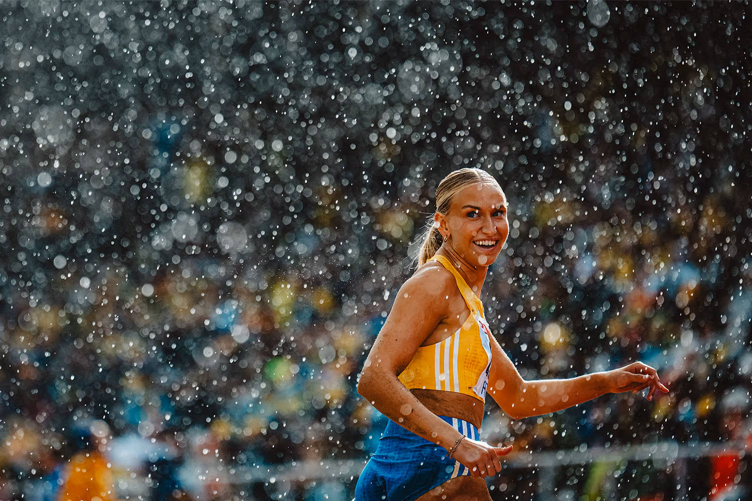Leende kvinnlig idrottare i gult och blått sportställ ler under regn i en stadion.