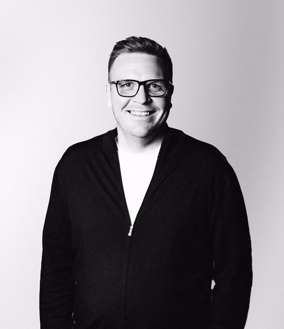 Leende man med glasögon, kort hår och mörk zip-up tröja över vit t-shirt, mot en ljus bakgrund.