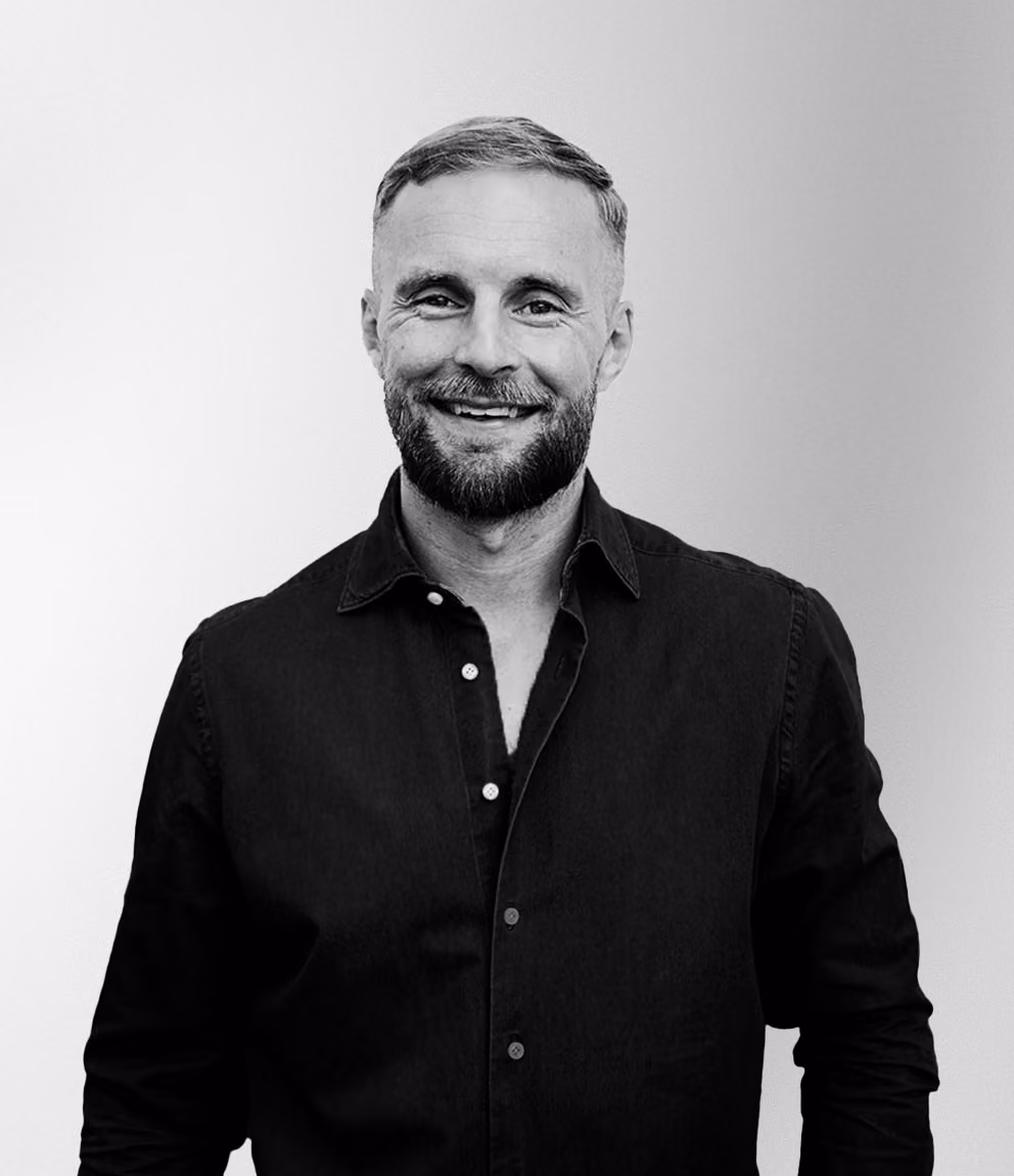 Porträtt i svartvitt av en leende man med kort hår och skägg, klädd i en mörk knappad skjorta mot en ljus bakgrund.
