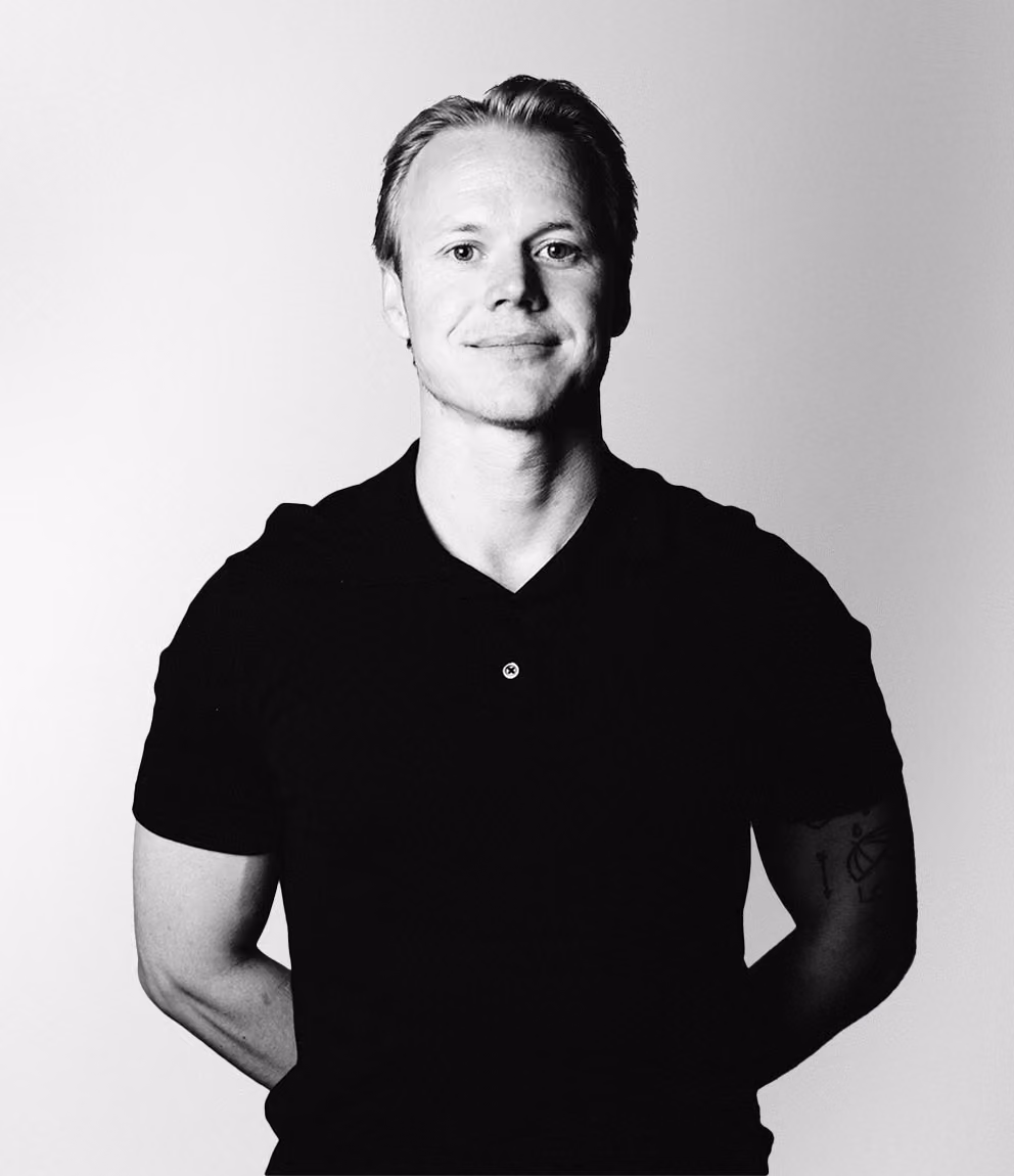 Ung man med ljust hår och svart pikétröja står mot ljus bakgrund med armarna bakom ryggen och ler lätt mot kameran.