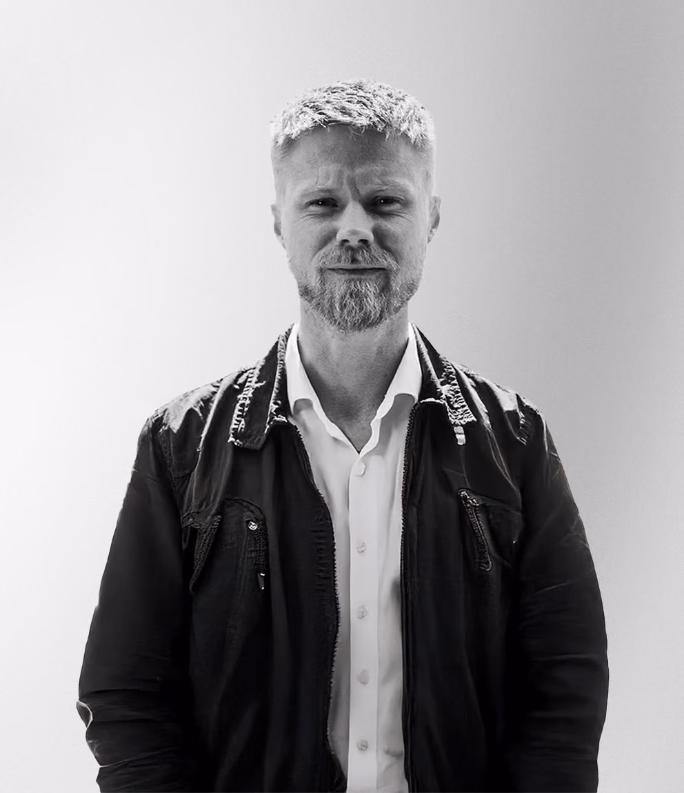 Porträtt i svartvitt på en man med kort ljust hår och skägg, klädd i vit skjorta och mörk jacka, mot en ljus bakgrund.