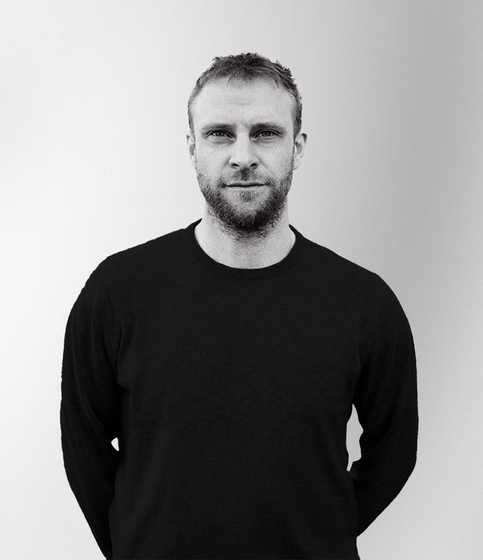 Porträtt i svartvitt av en man med kort ljust hår och skägg, iförd en svart tröja, stående mot en ljus bakgrund med händerna bakom ryggen.