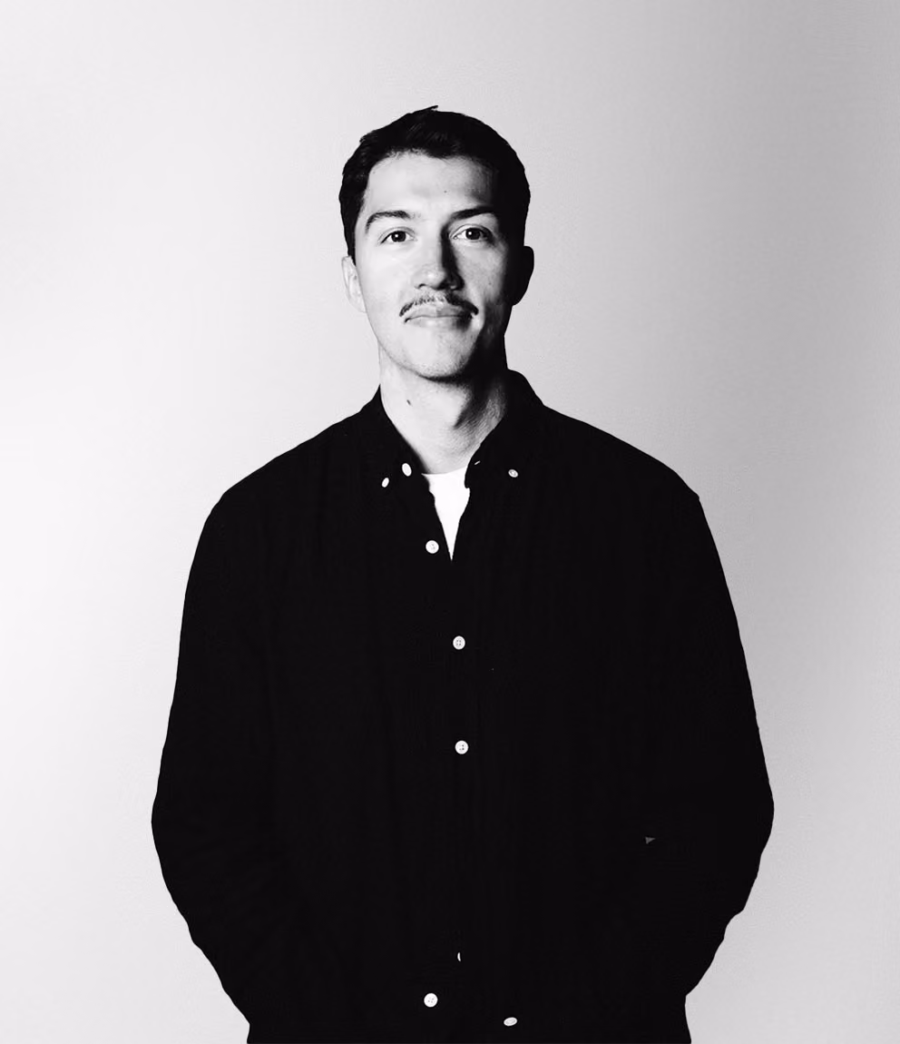 Svartvitt porträtt av en man med mustasch som bär svart skjorta över en vit t-shirt, står framför en ljus bakgrund och tittar rakt fram med lätt leende.