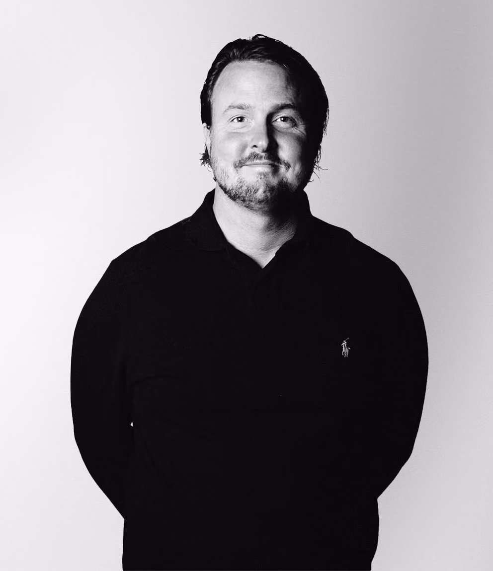 Man with short dark hair and a beard, wearing a black long-sleeve shirt with a small embroidered logo on the left chest, standing against a plain light background.