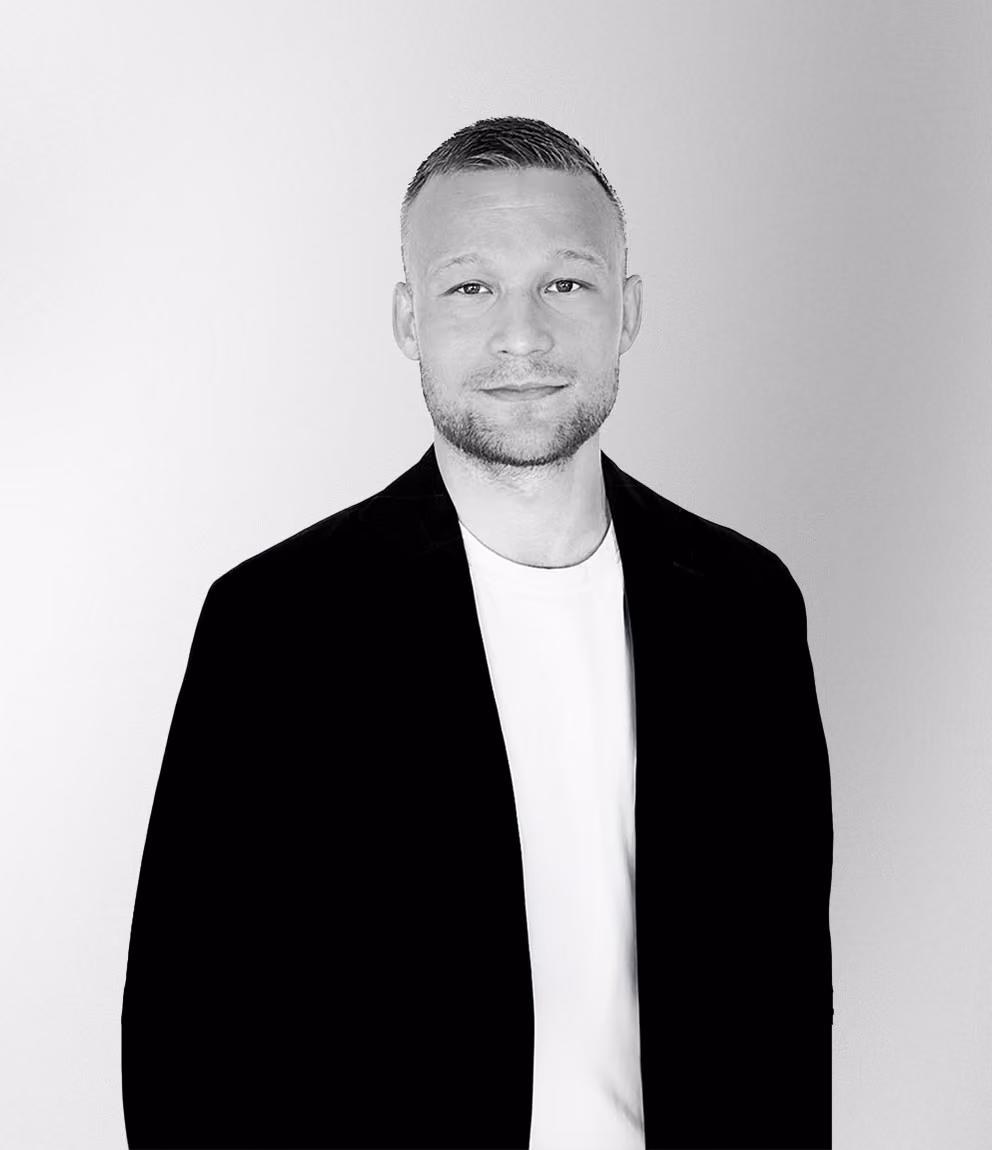 Porträtt i svartvitt av en man med kort hår och skäggstubb, iklädd svart kavaj och vit t-shirt, mot en ljus bakgrund.