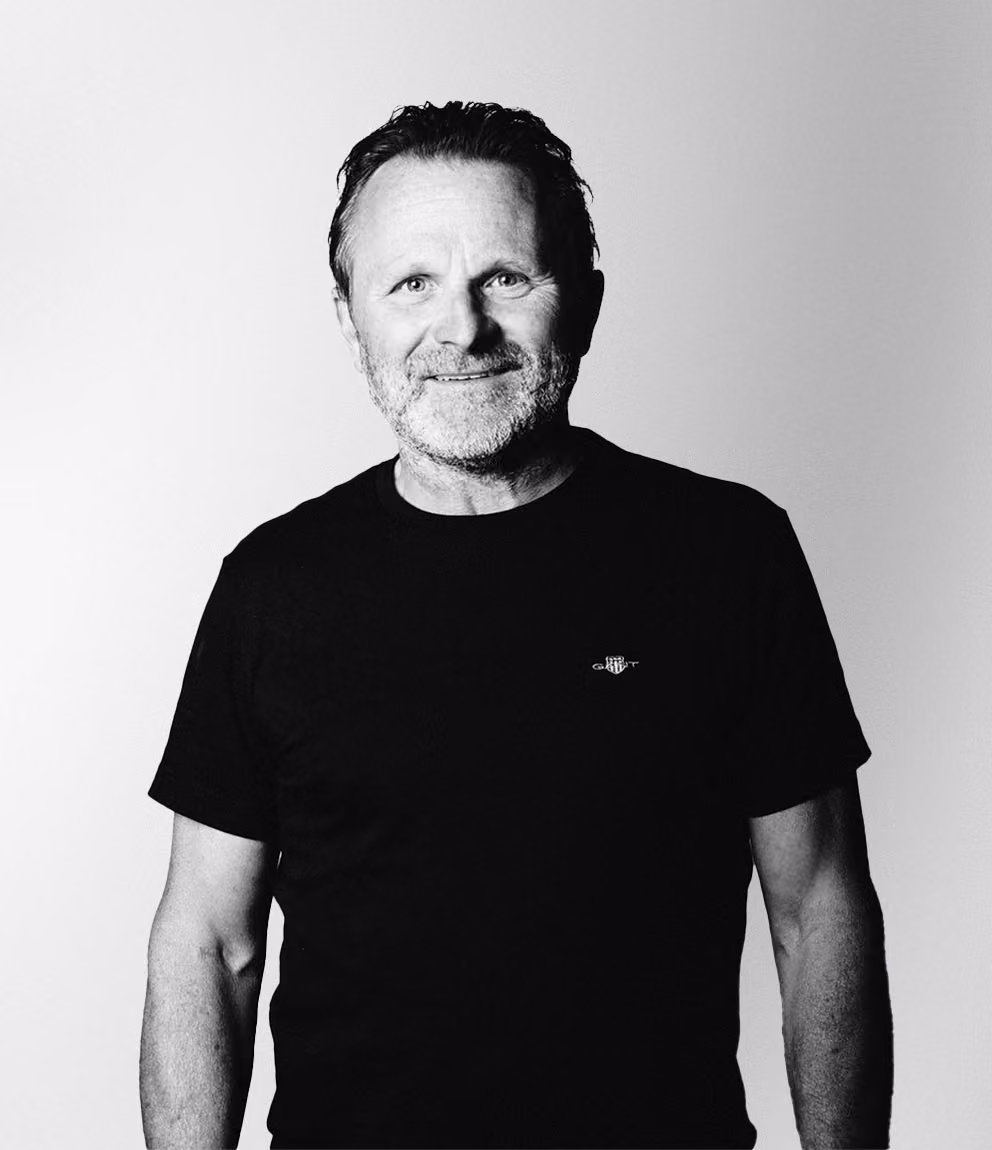 Porträtt i svartvitt av en leende medelålders man med kort hår och skägg, klädd i en svart t-shirt med liten logga på bröstet, mot en ljus bakgrund.