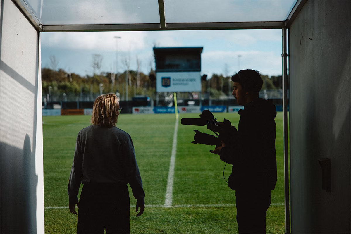 Två personer står i en tunnel som leder ut till en grön fotbollsplan, den ena håller en professionell videokamera.