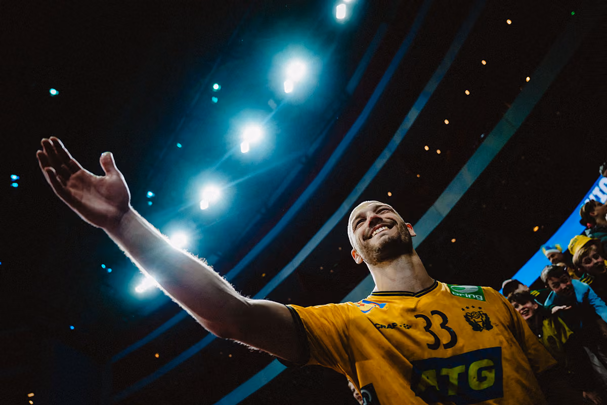 En handbollsspelare i gul tröja med nummer 33 ler och sträcker ut armen mot publiken i en upplyst arena.