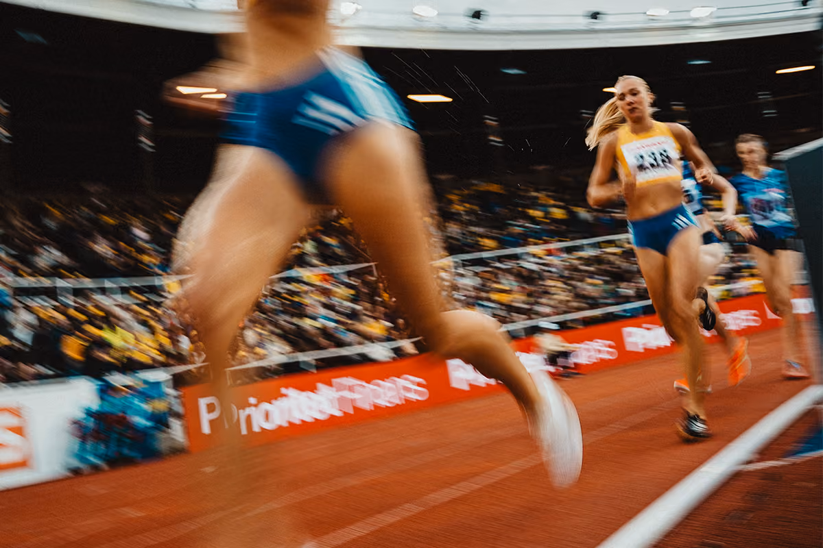 Snabbt rörelseoskärpefoto av kvinnliga löpare i blå sportkläder som tävlar på en löparbana inomhus med en publik i bakgrunden.
