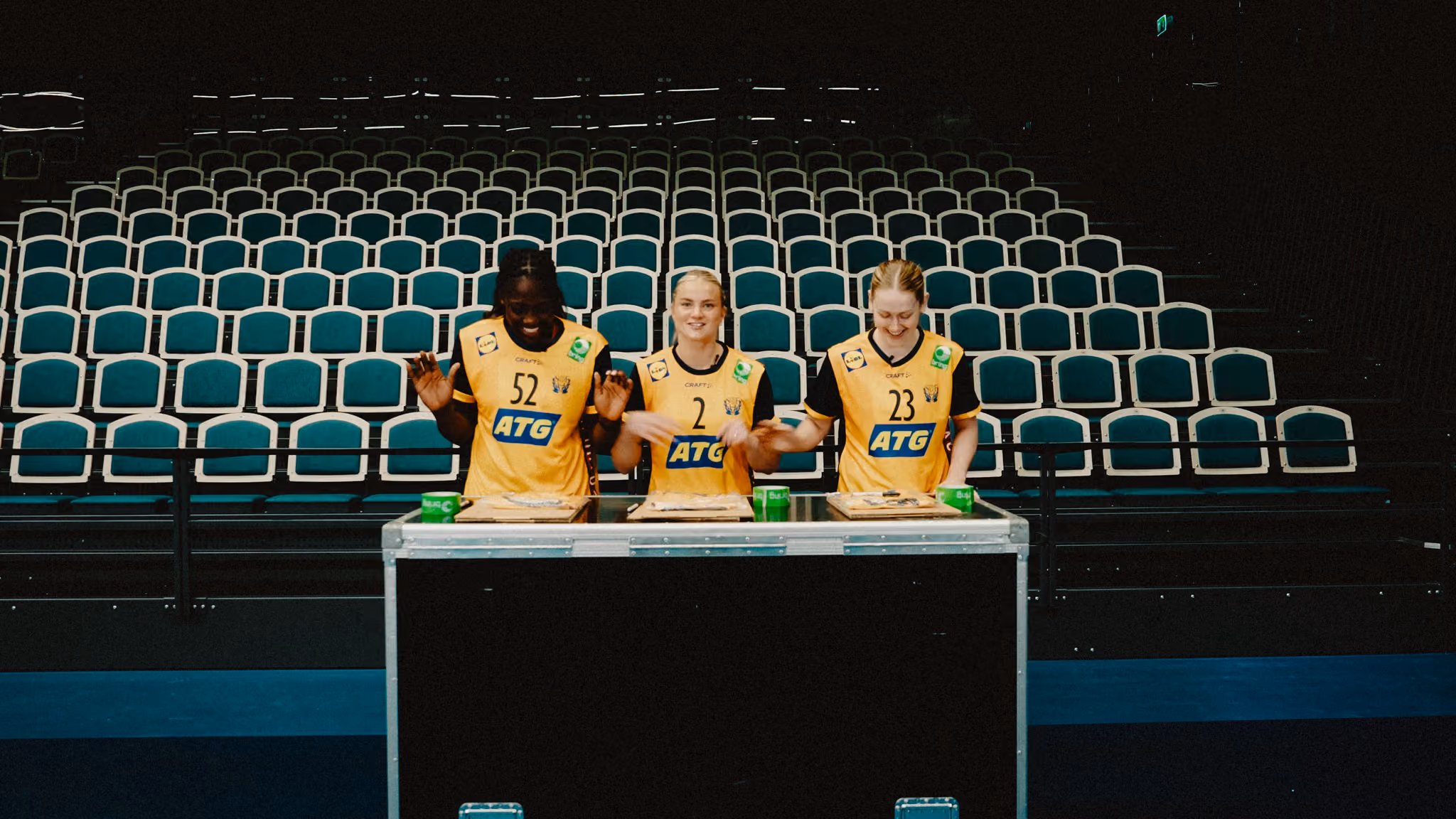 Tre handbollsspelare i gula och svarta matchtröjor står bakom en svart bänk i en nästan tom arena med gröna och vita sittplatser.