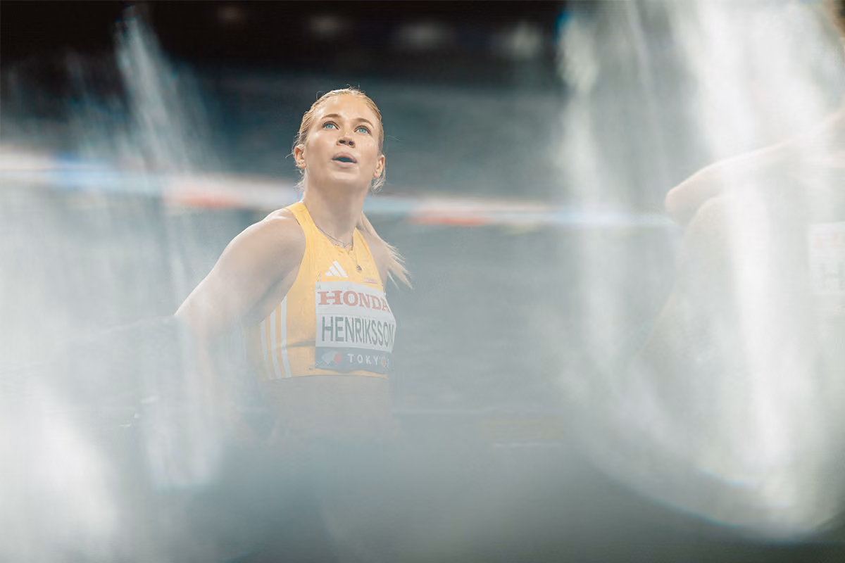 Kvinna idrottare med blont hår i hästsvans iklädd gul sporttop med nummerlapp och texten Honda och Henriksson tittar uppåt i en dimmig eller suddig miljö.