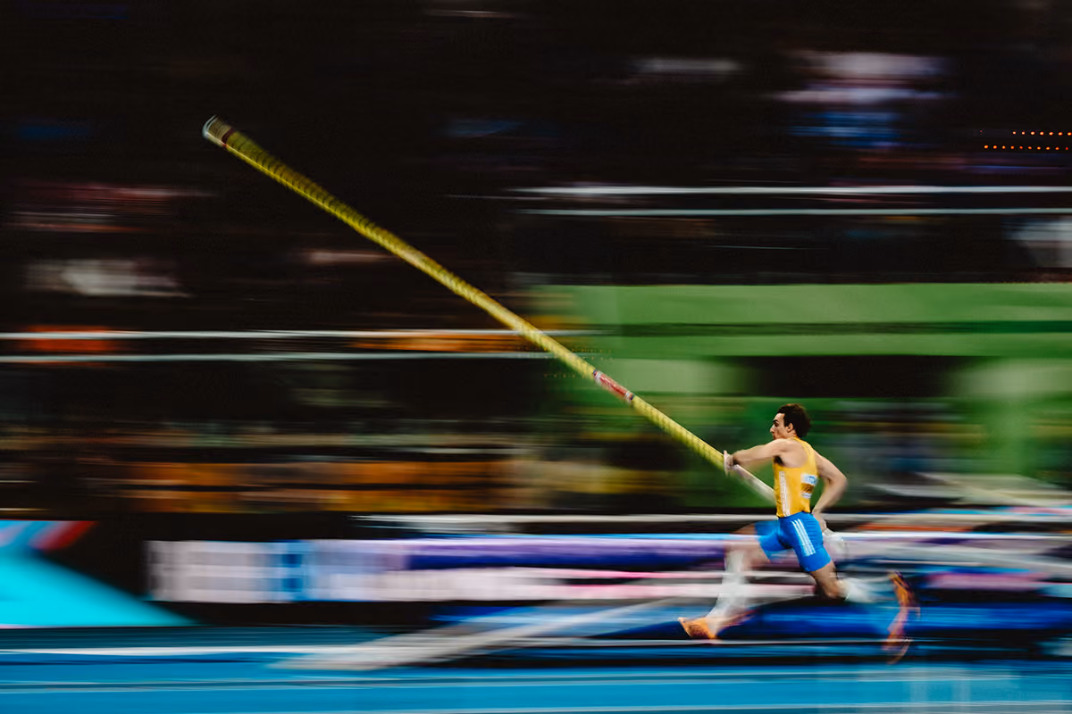 En stavhoppsidrottare i blå shorts och gul tröja springer med staven mot hoppmattan i en idrottsarena.