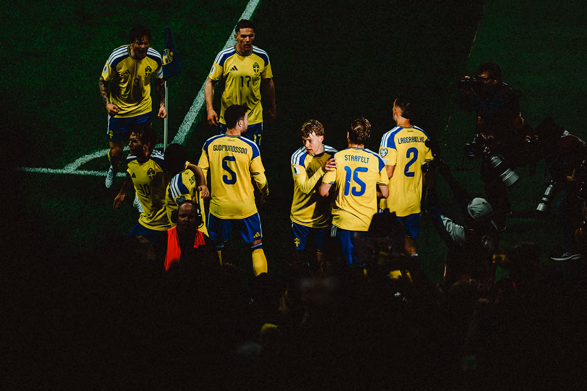 Svenska fotbollsspelare i gula tröjor och blå shorts samlas nära hörnflaggan på en gräsplan, fotografer och publik syns i förgrunden.