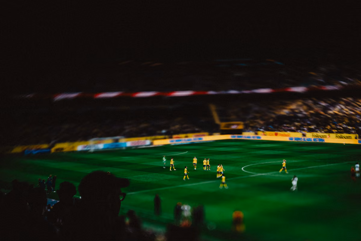 Fotbollsmatch på kvällen med spelare i gult på den gröna planen och suddiga åskådare i förgrunden.