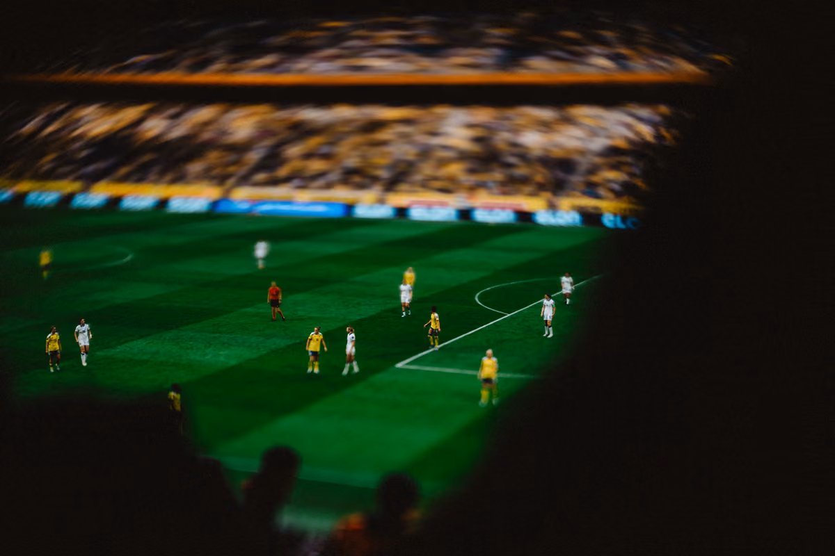 Fotbollsmatch sedd från läktaren med spelare i gula och vita tröjor på grön plan och suddiga åskådare i förgrunden.
