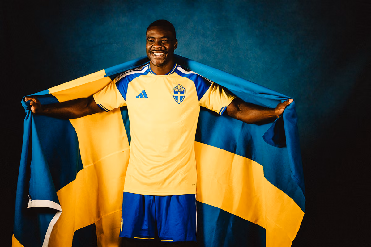 Glad man bär en svensk fotbollströja i gult och blått och håller en stor svensk flagga utspänd bakom sig mot en mörk bakgrund.