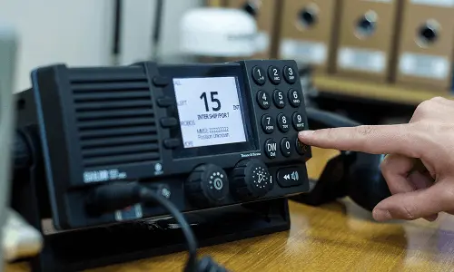 A hand pressing a button on a black office telephone with a digital screen showing the date and time.