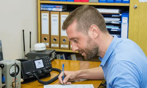 Man in blue shirt writing on a sheet of paper at a desk with radio communication equipment.