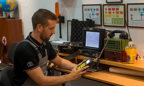 Man in black shirt using a handheld radio device connected to a monitor on a desk with technical equipment.