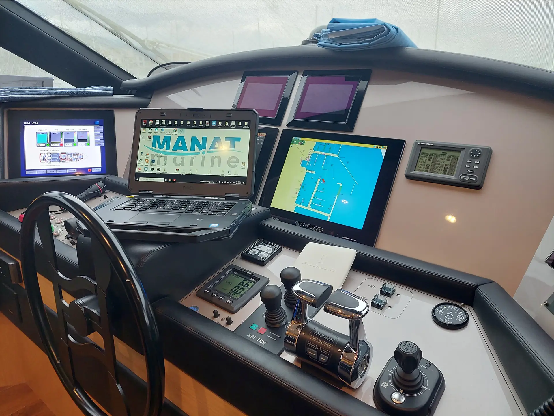 Boat control panel with steering wheel, laptop displaying MANAT marine logo, multiple monitors showing navigation and system data, and various control levers and buttons.