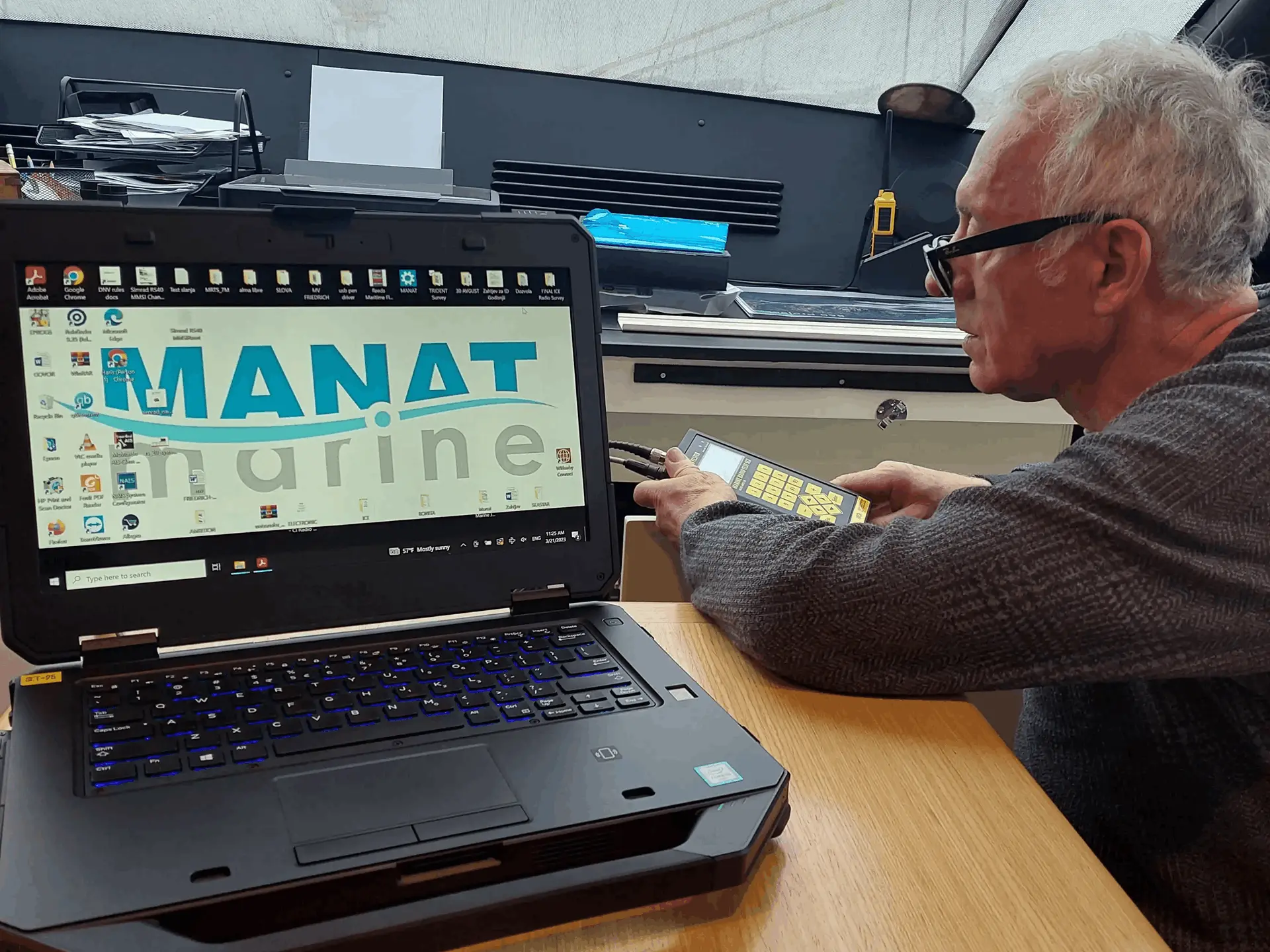 Older man wearing glasses operating a handheld device beside a laptop displaying 'MANAT marine' on the screen.