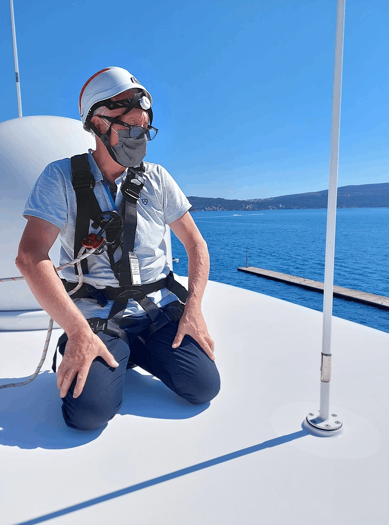 Man wearing safety harness, helmet, glasses, and face mask kneeling on a white boat deck with blue sea and sky in the background.