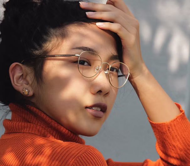 Young person wearing glasses and orange turtleneck sweater, touching hair outdoors.