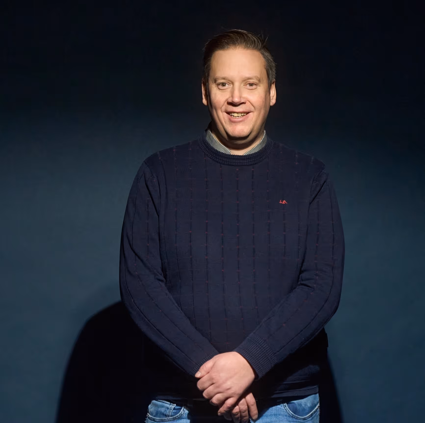 Mann med mørk genser og lyse jeans smiler foran en mørk bakgrunn.