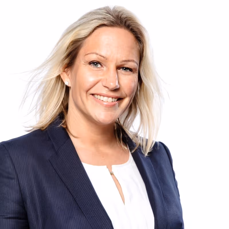 Smilende kvinne med blondt hår i mørk blazer og hvit topp foran hvit bakgrunn.