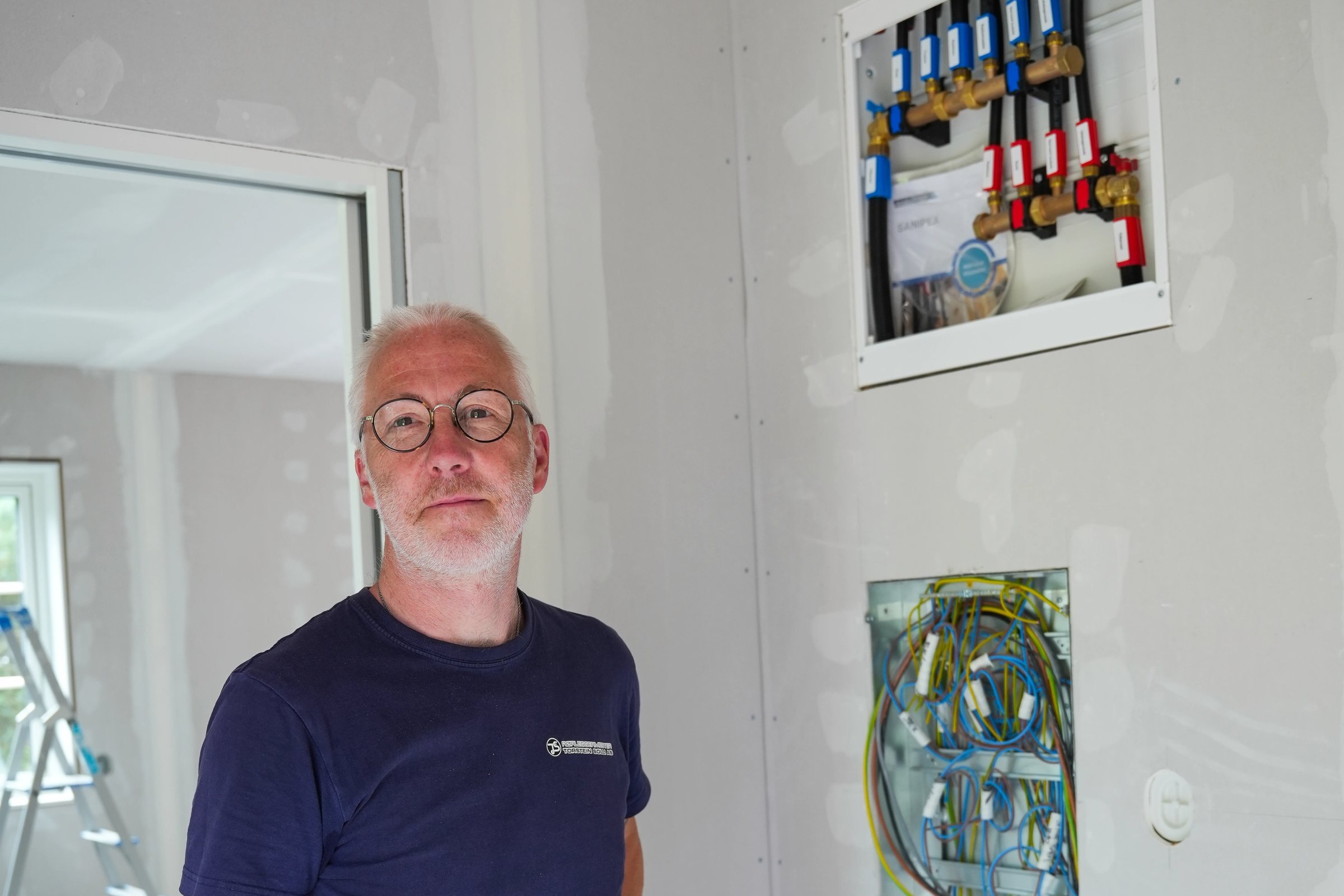 Mann med briller og blå t-skjorte foran elektrisk koblingsskap i byggeplass.