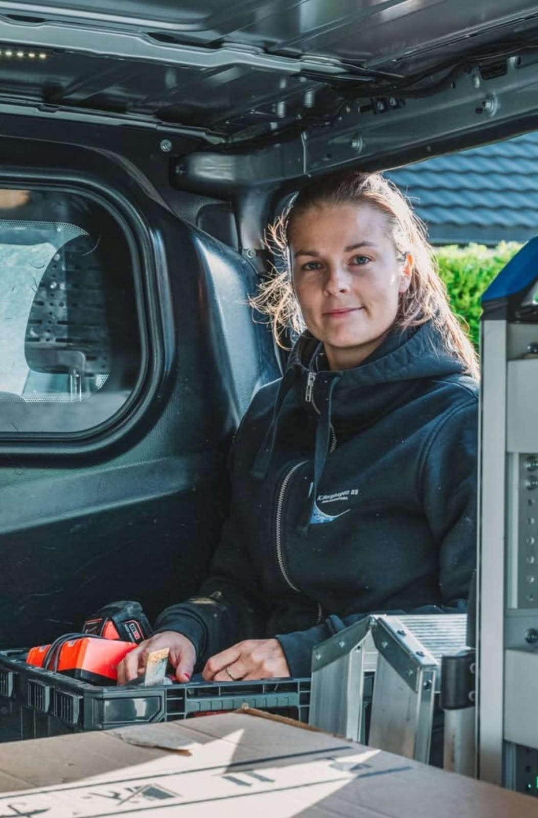 Kvinne i mørk jakke sitter i varebil med verktøy og esker rundt seg.