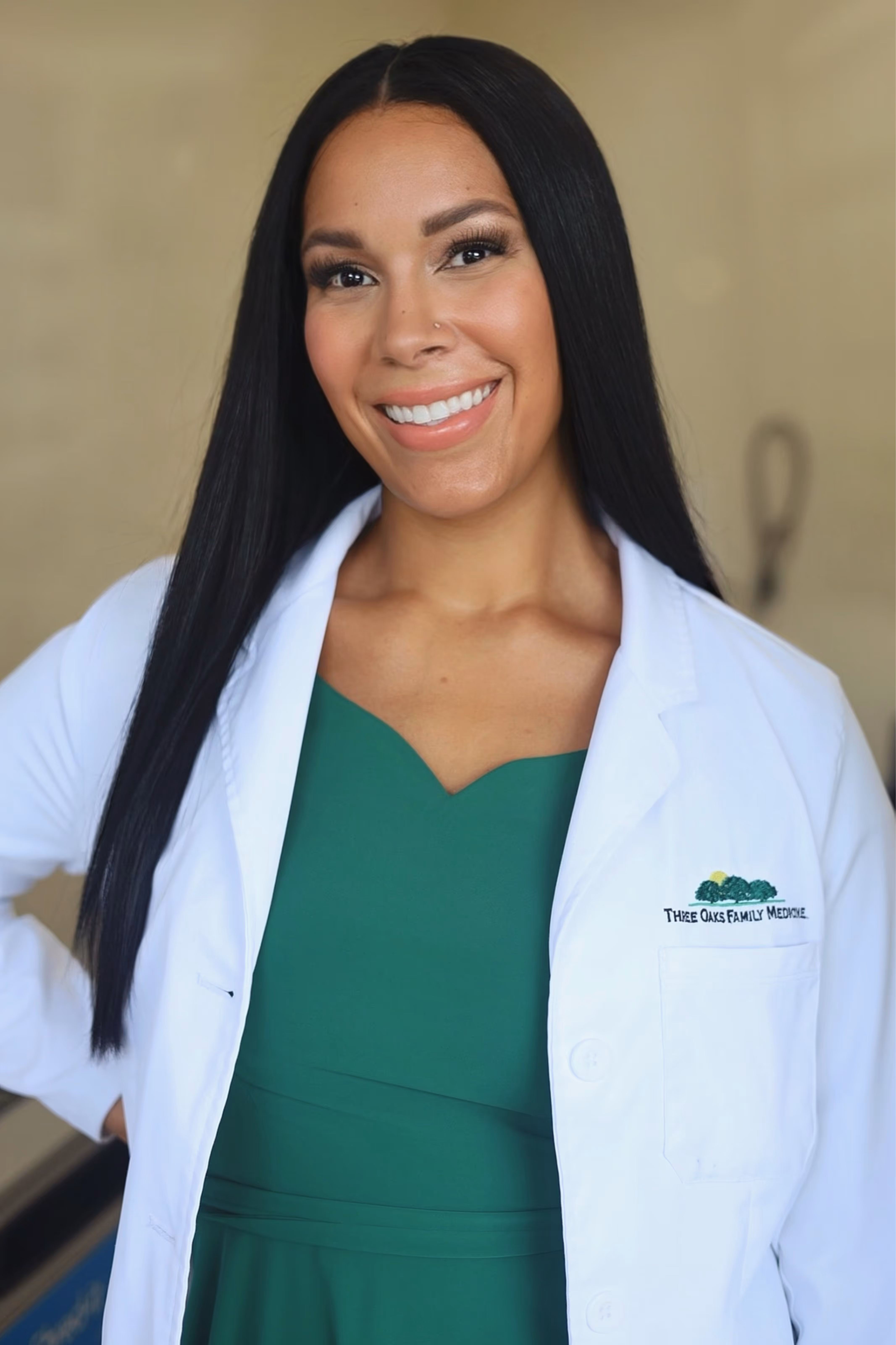 Smiling woman with long black hair wearing a green dress and white medical coat with Three Oaks Family Medicine logo.