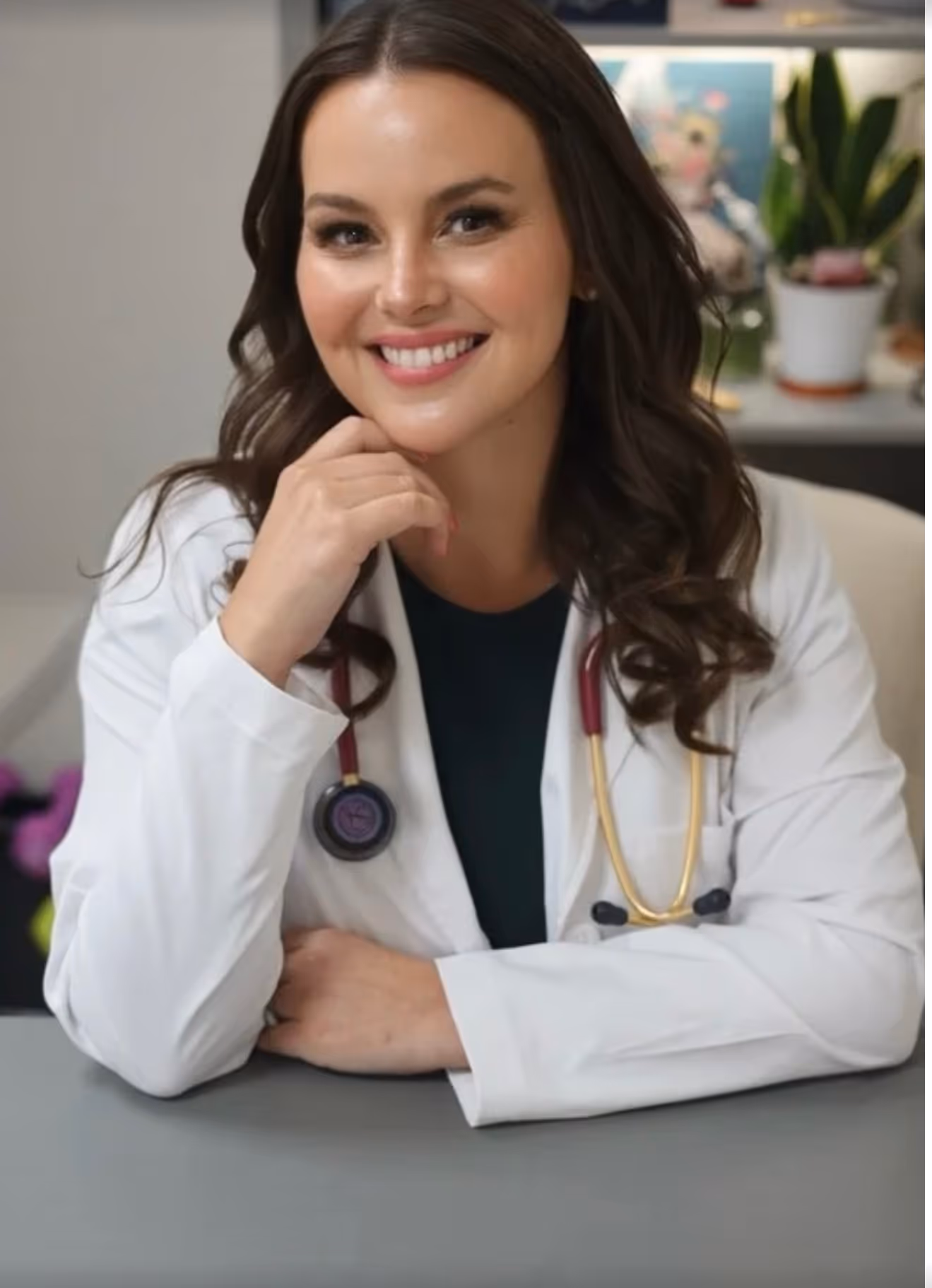 Smiling female doctor with long dark hair wearing a white coat and stethoscope sitting at a desk.