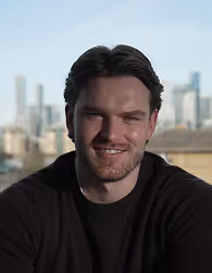 Smiling man with dark hair and beard wearing a black shirt, with a cityscape background.