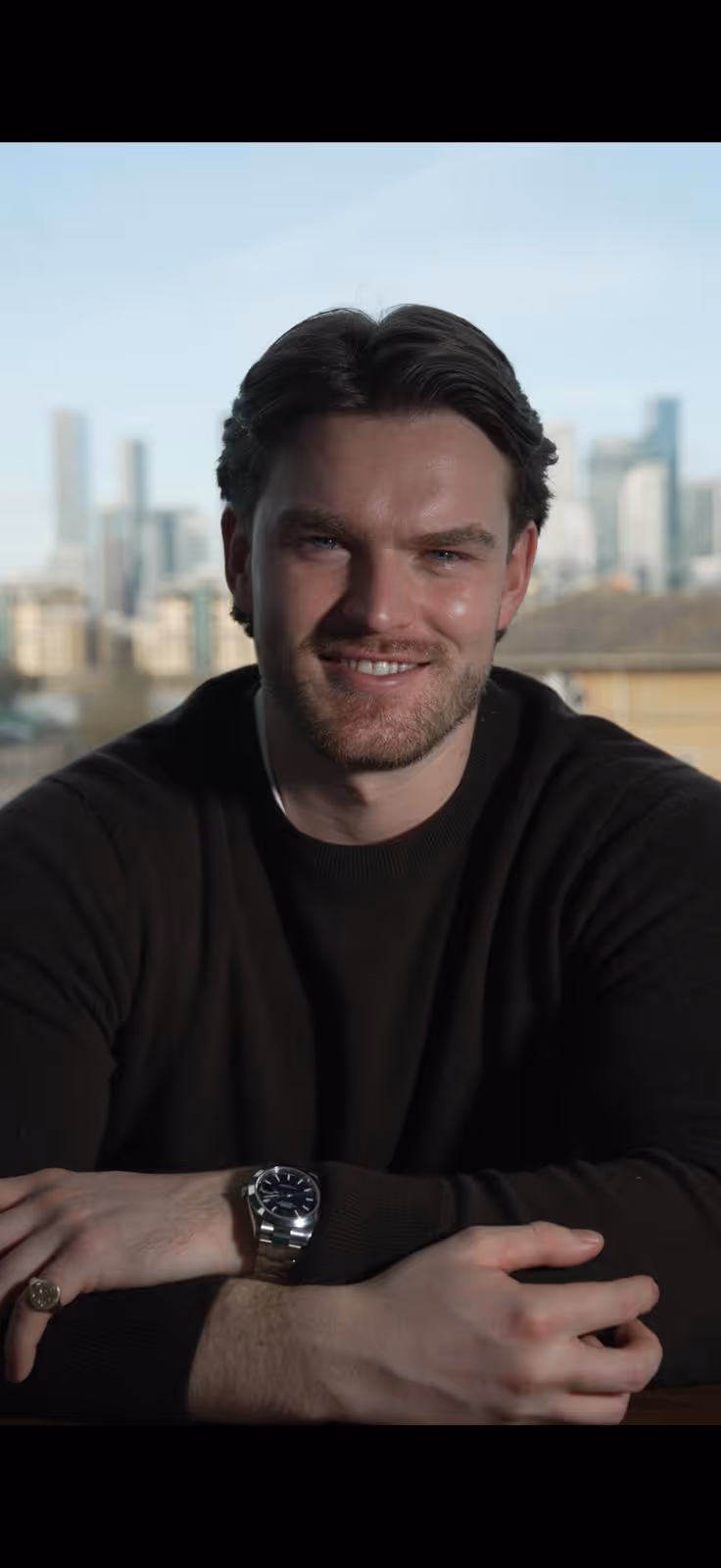 Smiling man with dark hair and beard wearing a black sweater and a silver wristwatch against a city skyline background.