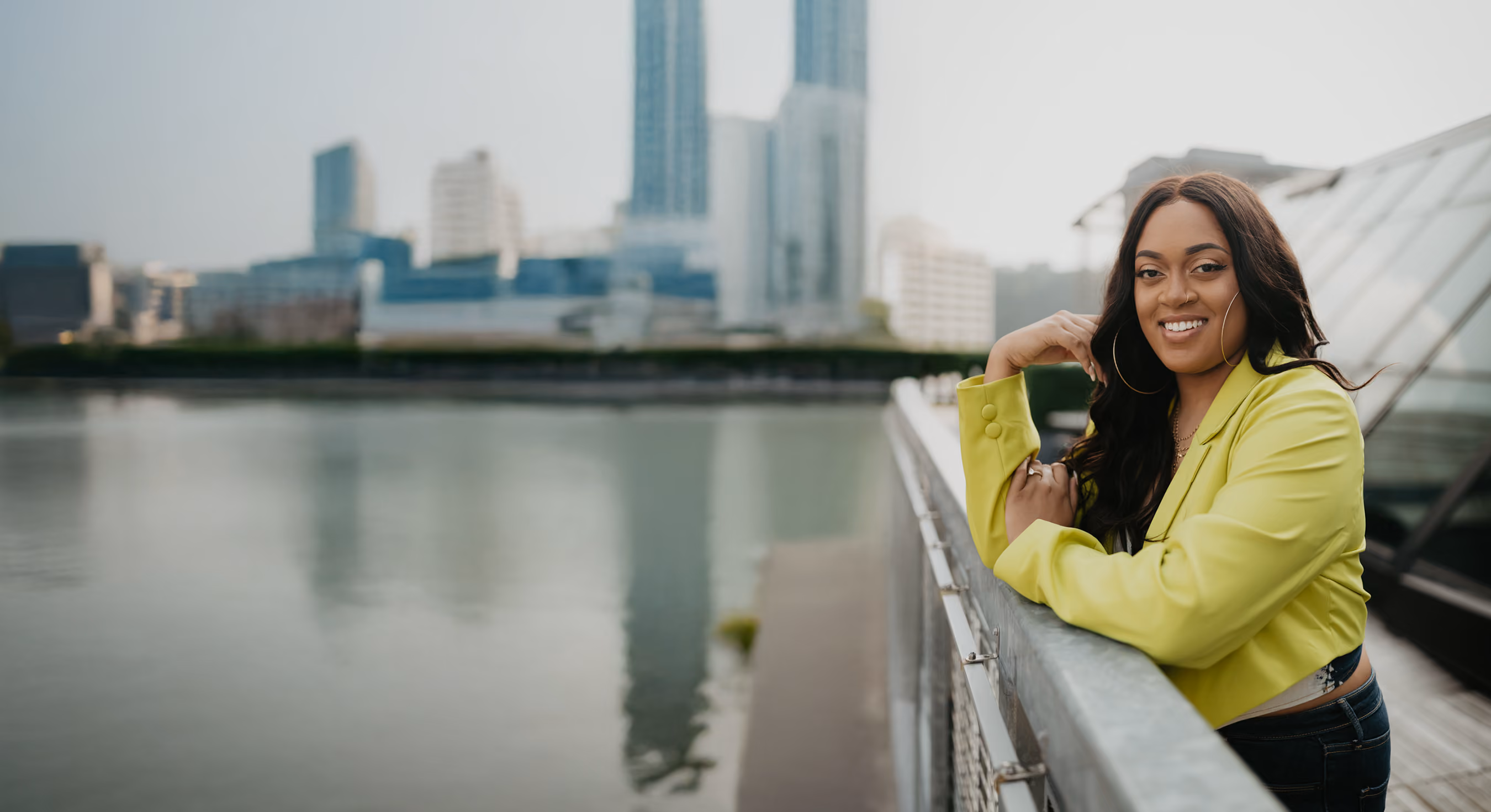 Pittsburgh website designer Domanique Taylor leaning on a railing by the river with the city skyline behind her