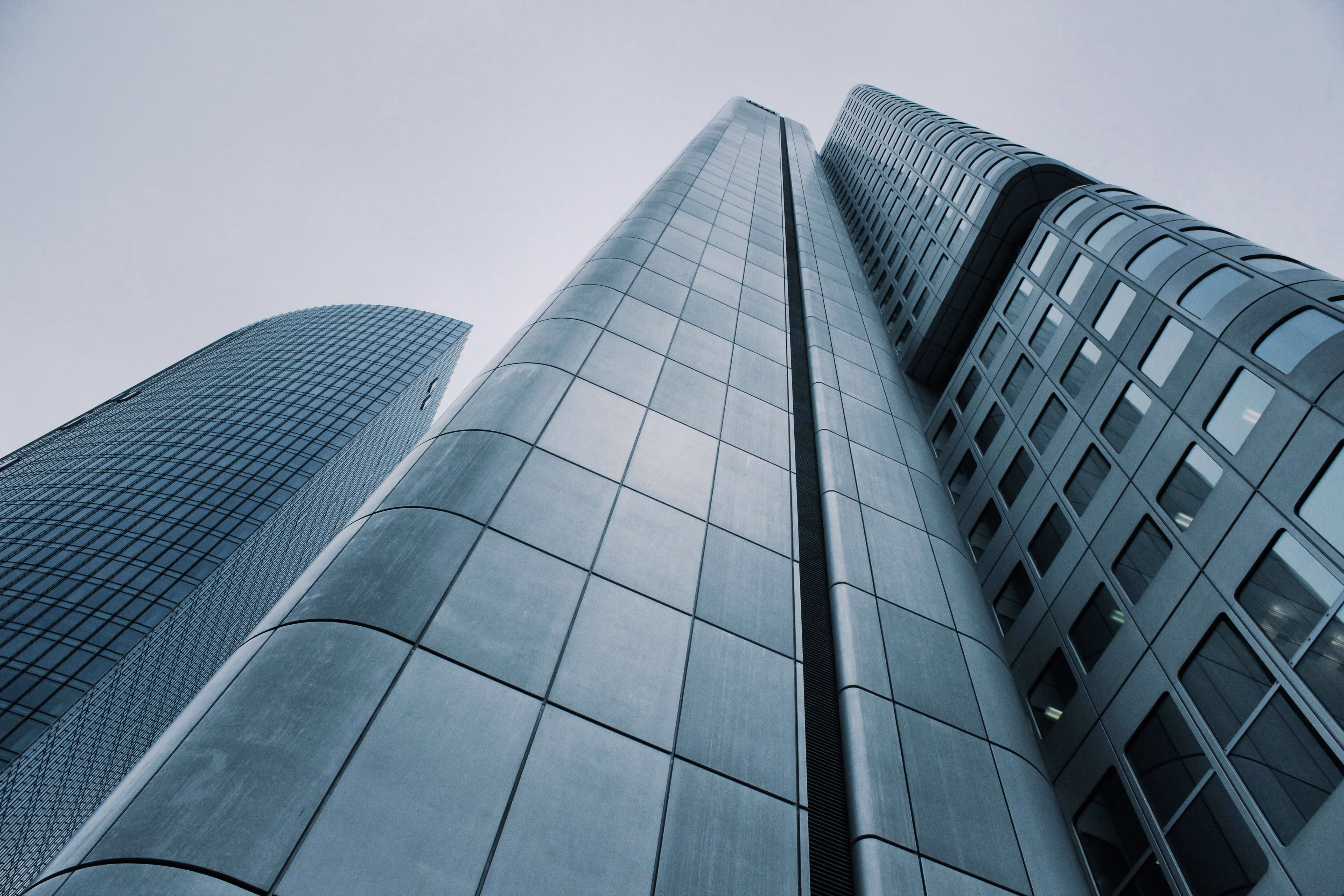 Looking up at three modern skyscrapers with glass and metal facades against a gray sky.