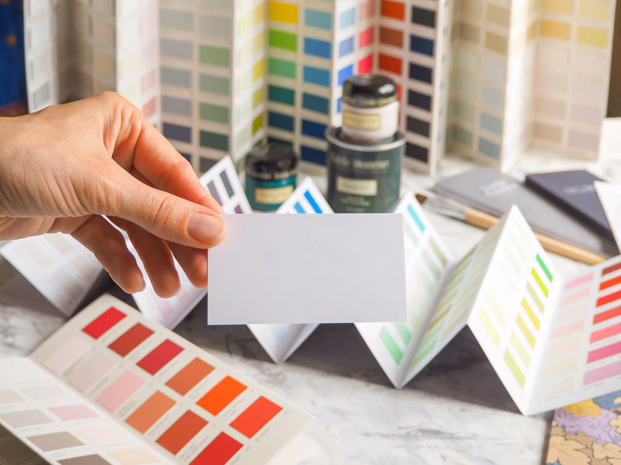Mão segurando uma amostra de cor branca à frente de vários catálogos de cores dispostos em leque sobre uma mesa.
