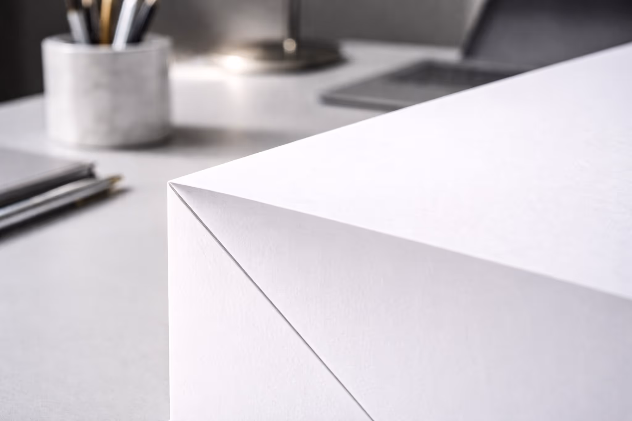 Plano de uma mesa branca com uma caixa retangular branca em foco no primeiro plano e vários itens fora de foco ao fundo, incluindo um suporte para canetas e canetas.