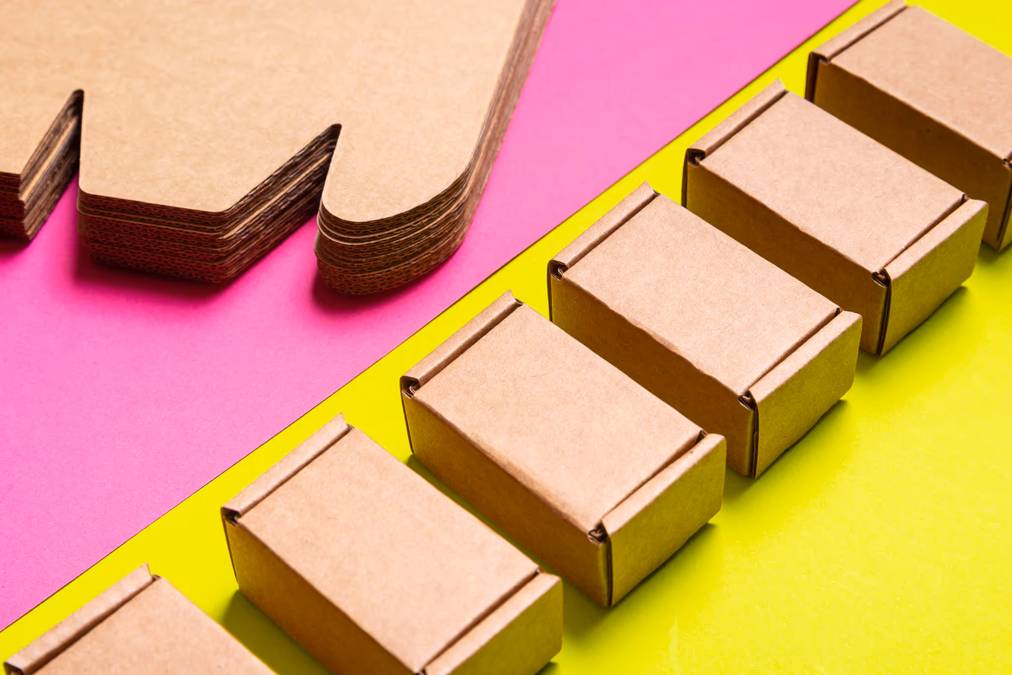 Caixas de cartão empilhadas alinhadas em fundo amarelo e chapas de cartão ondulado em fundo rosa.