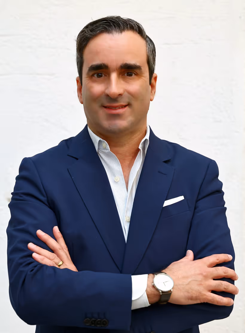 Homem de cabelo curto e escuro vestindo blazer azul com camisa branca, braços cruzados e sorrindo diante de fundo branco.