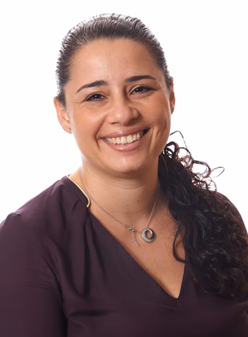 Mulher sorridente com cabelo preso para trás, vestindo blusa roxa e colar prateado em fundo branco.