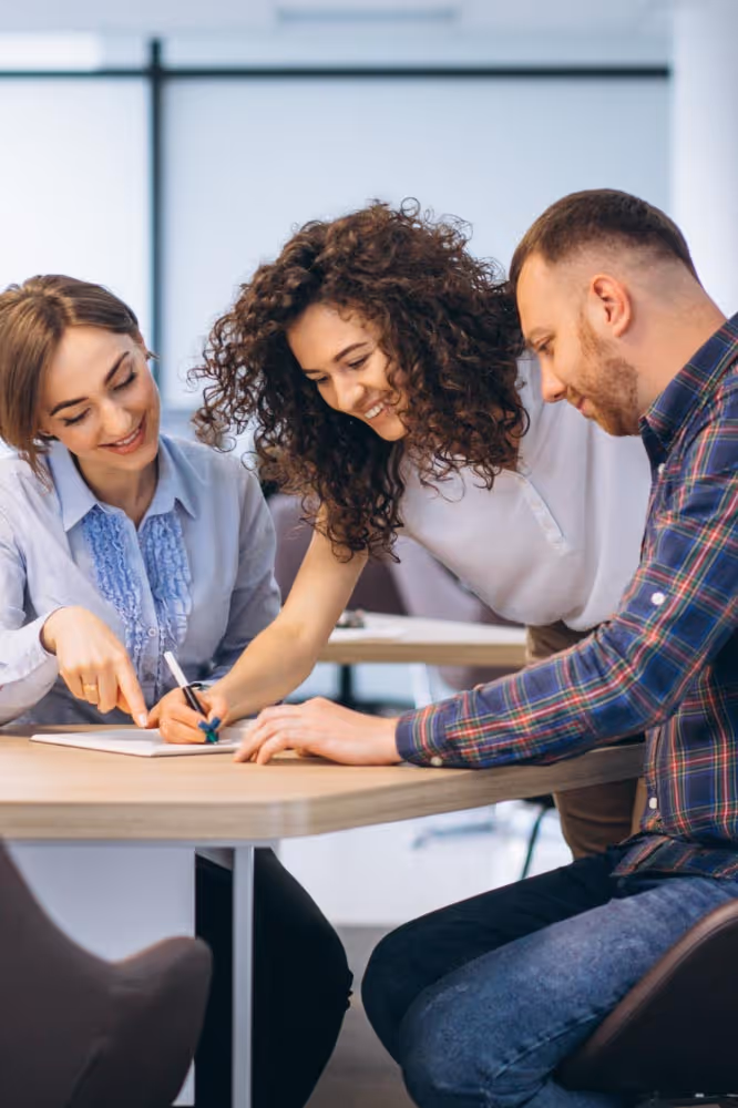 Três pessoas colaboram numa mesa, uma escreve enquanto outra aponta e sorri, num ambiente de escritório moderno.