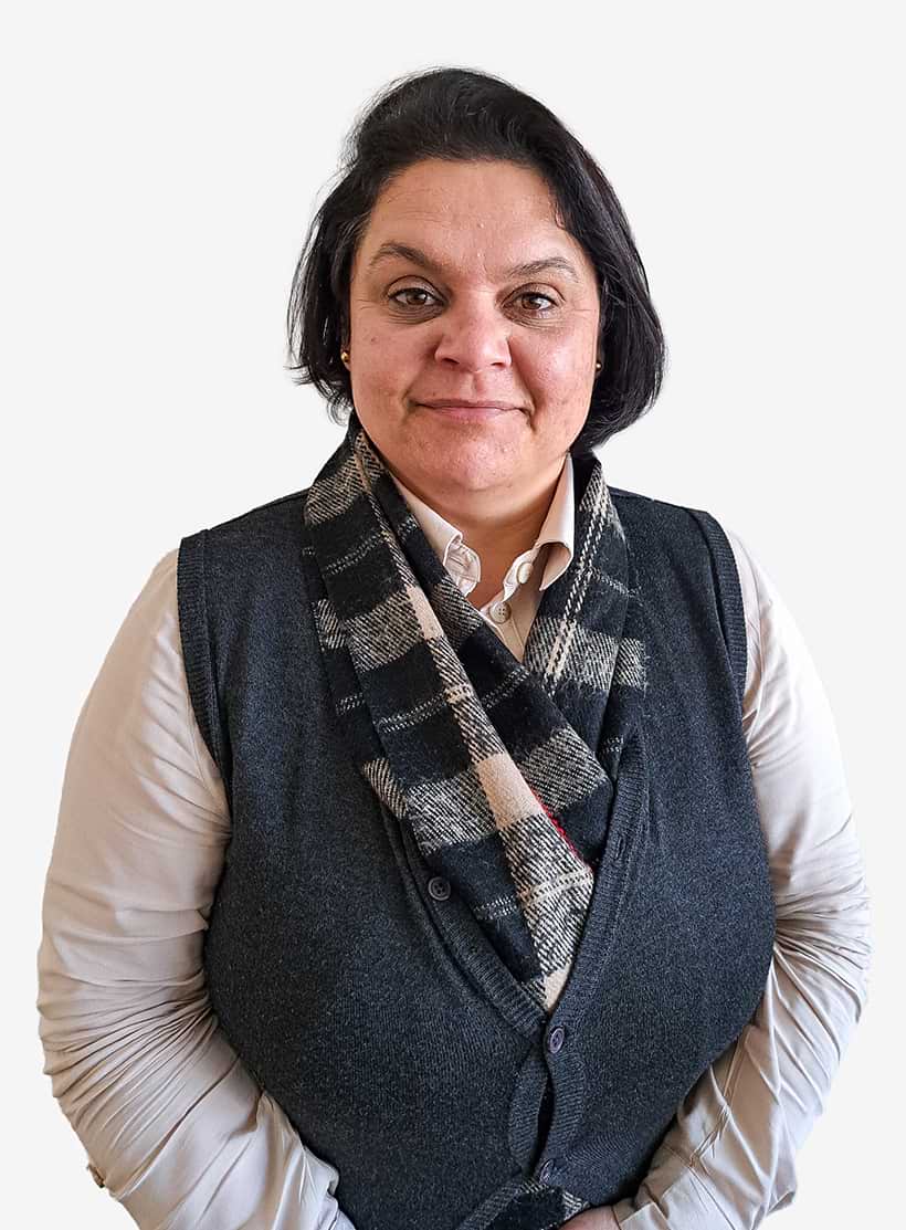 Mulher com cabelo preto curto, vestindo camisa clara, colete cinza escuro e cachecol xadrez preto e branco, posando com as mãos cruzadas.