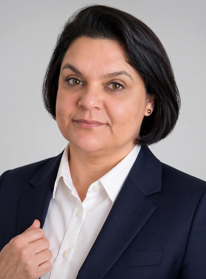 Mulher adulta veste camisa branca e blazer azul escuro, posa com expressão neutra e cabelo preto curto.
