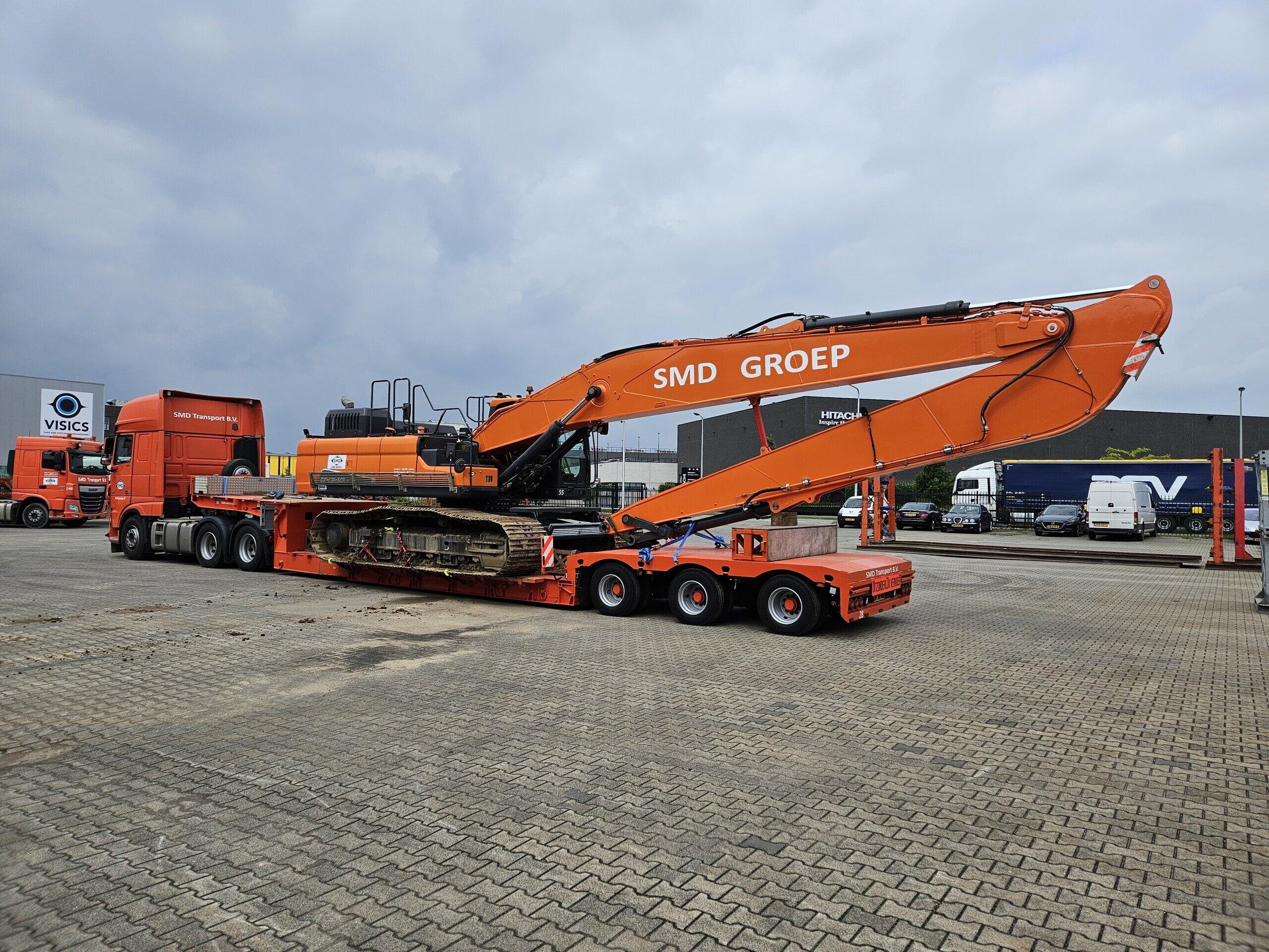 Oranje graafmachine met 'SMD GROEP' op de arm geladen op een vrachtwagentrailer op een parkeerterrein bij een bedrijf.